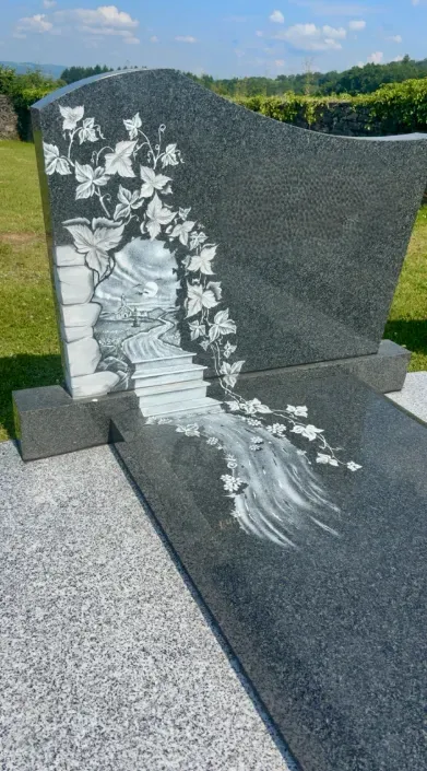 Stèle funéraire personnalisée en granit foncé gravée d'un paysage et de lierre.